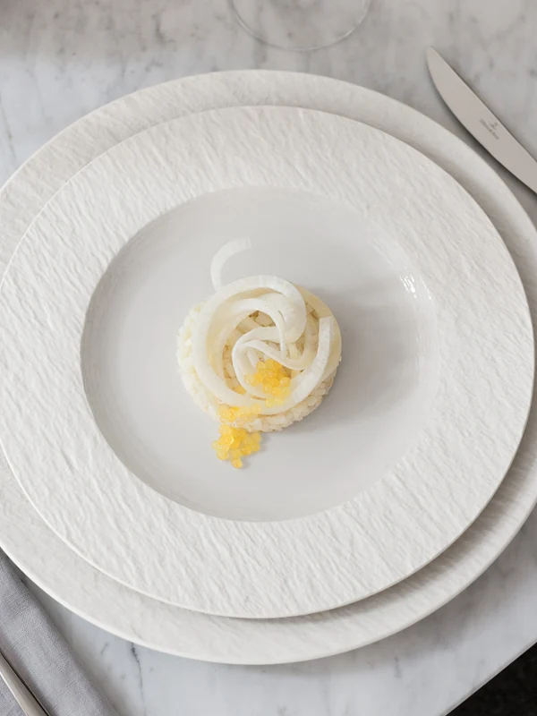 A small portion of food on a white Manufacture plate A small portion of food is arranged on a white Manufacture plate with a knife, fork, and napkin on a marble surface.