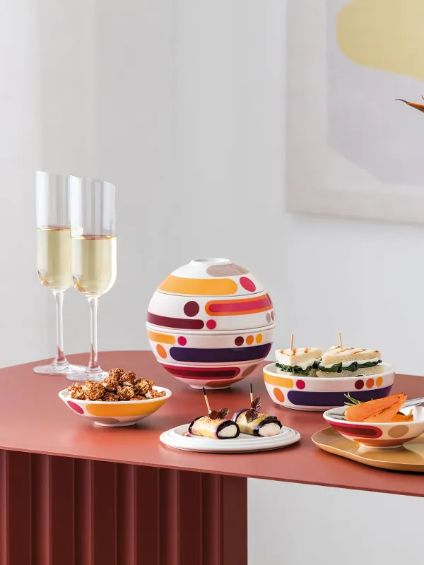 Tavola moderna con La Boule Miami, assortimento di snack, due calici da champagne e un vaso bianco Manufacture Collier con fiori arancioni.