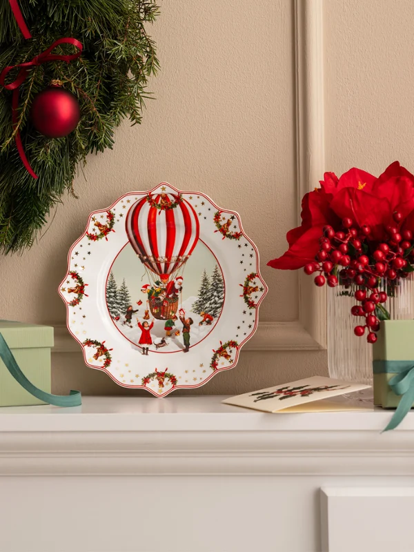 A Christmas wreath, a decorative plate featuring a hot-air balloon by Villeroy & Boch, green gift boxes, and red flowers, arranged on a white mantelpiece.