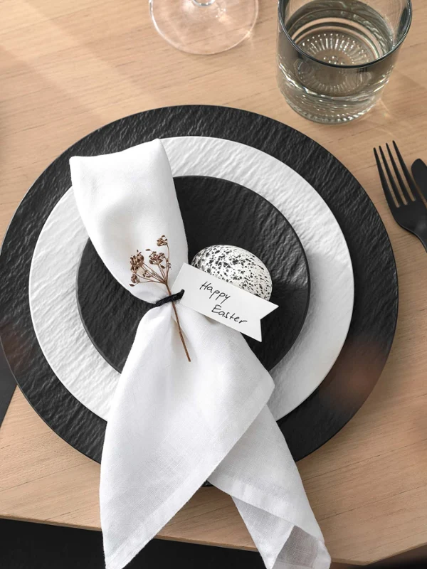 An elegant Easter table setting with Manufacture crockery and cutlery as well as a decorative napkin and a personalised note on a wooden table.