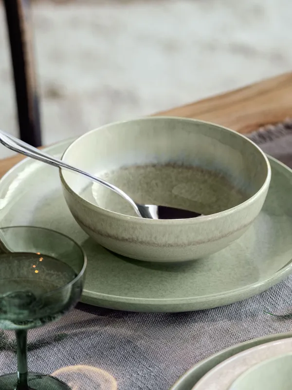 Un bol Perlemor vert avec une cuillère repose sur une assiette assortie, posée sur une table avec des bougies et des décorations végétales