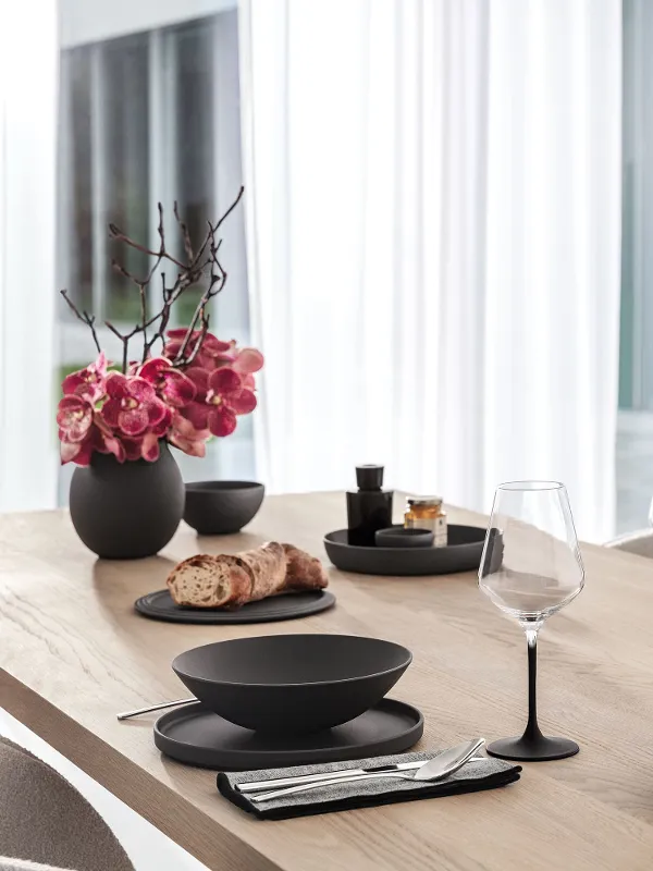 A wooden dining table set with a Villeroy & Boch black La Boule, Manufacture glasses and Piemont cutlery.