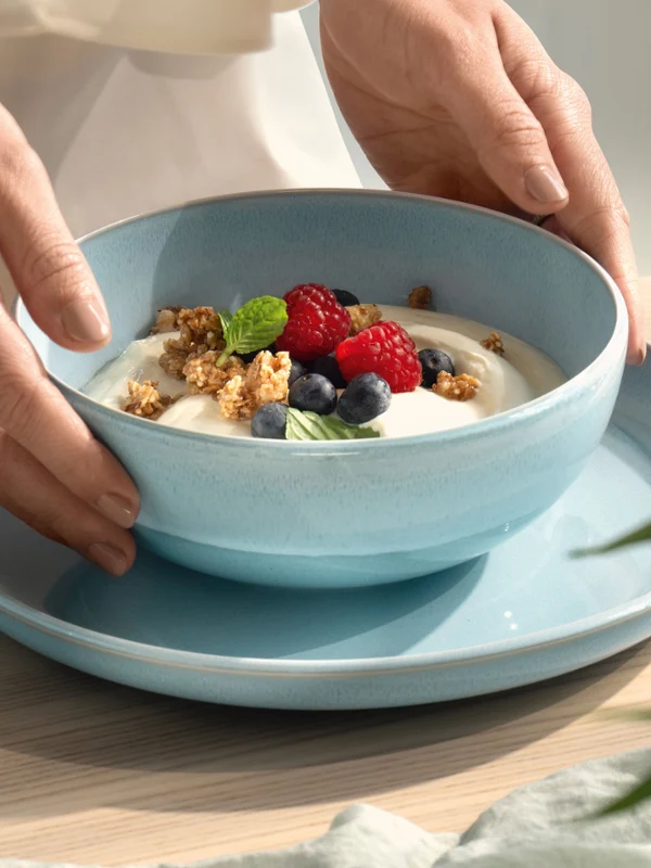 Hands placing a blue mother-of-pearl aqua bowl with yoghurt on a blue plate Hands placing a blue mother-of-pearl aqua bowl with yoghurt, muesli, blueberries and raspberries on a blue plate.