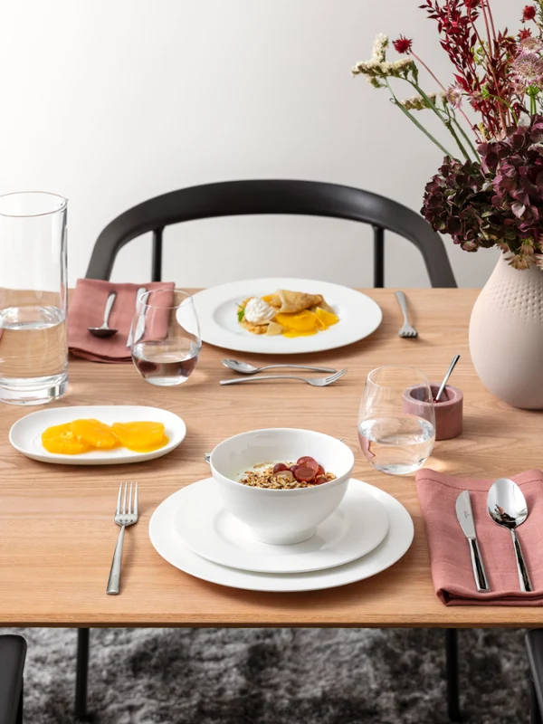 A wooden dining table with plates full of mango slices, a dish of poached egg and a bowl of muesli, plus glasses, cutlery and a vase of flowers.	