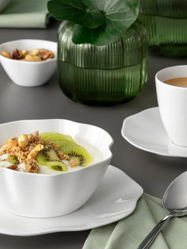 Eine weiße Pura Schale mit Müsli und Kiwi und eine Tasse Kaffee auf einer Untertasse Eine weiße Pura Schale mit Müsli und Kiwi und eine Tasse Kaffee auf einer Untertasse auf einem grauen Tisch mit einer grünen Serviette.