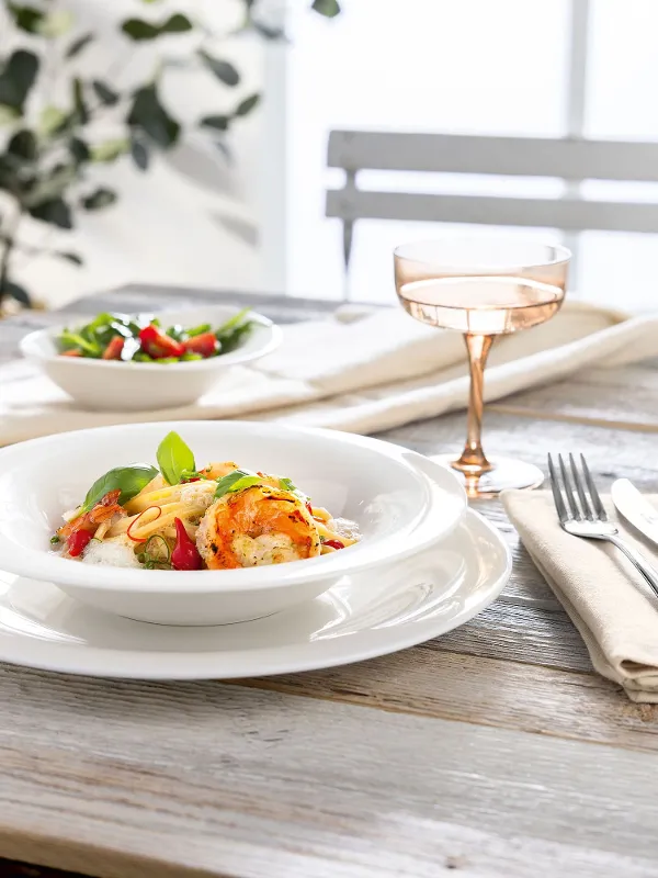 Un piatto di pasta con verdure ed erbe, un'insalata e una bevanda su un tavolo di legno apparecchiato per una persona.