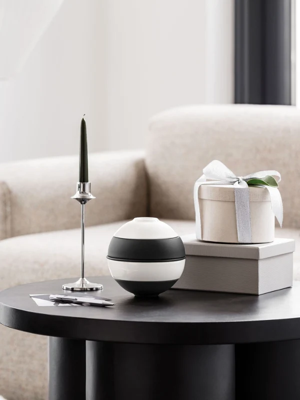 A round black coffee table with the Villeroy & Boch La petite Boule A round black coffee table with the Villeroy & Boch La petite Boule, a green candle on a silver holder and a gift box on top of two books. Beige sofa and a grey plinth with a vase in the background.