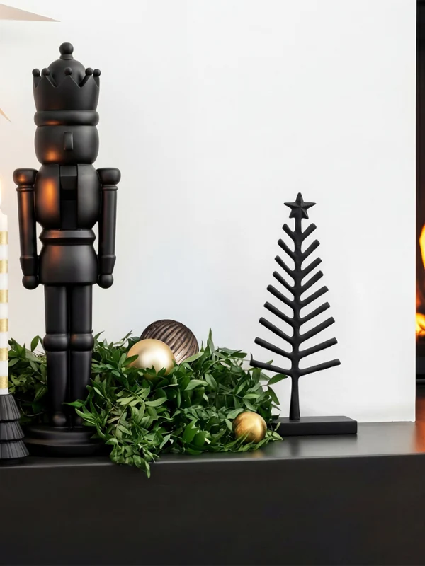 Un cascanueces negro, una vela y un árbol de metal se encuentran sobre una repisa de chimenea decorada, junto a una chimenea encendida.
