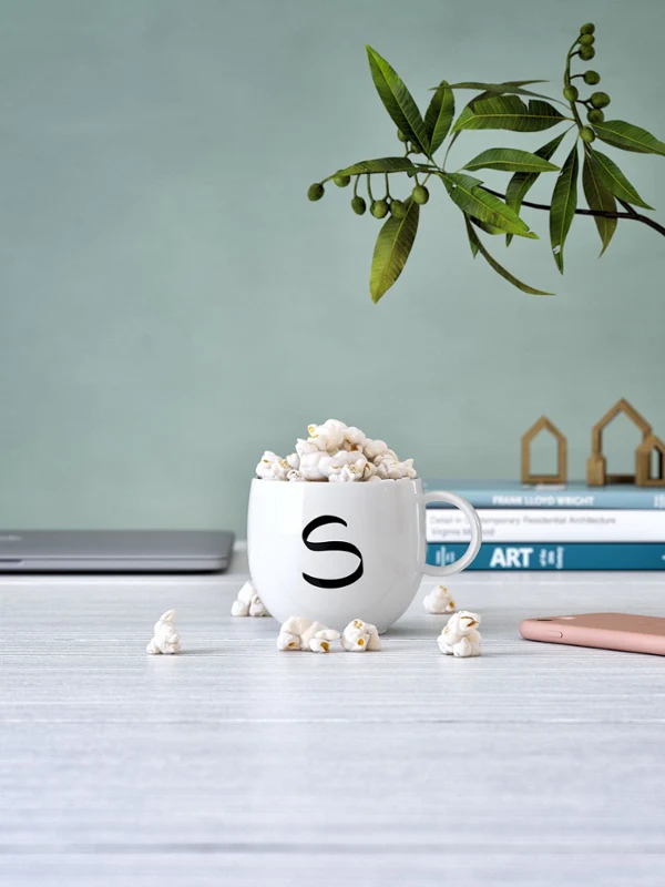 Kubek Letters z literą „s” wypełniony popcornem Kubek Letters firmy Villeroy & Boch z literą „s”, wypełniony popcornem, na biurku z laptopem, książkami i smartfonem w pobliżu.