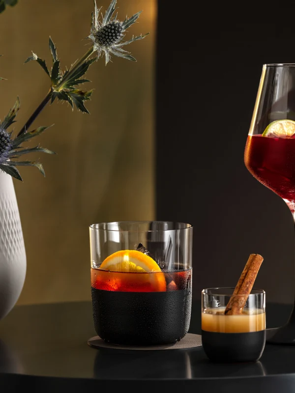 Twee cocktails in Manufacture glazen en een Manufacture vaas met een plant op een zwarte tafel tegen een gouden achtergrond.