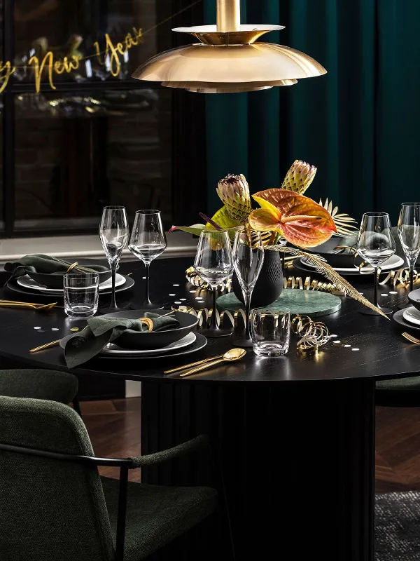 A round black dining table is elegantly set with green napkins, gold cutlery and a flower arrangement. Green chairs surround the table and a "Happy New Year" sign welcomes guests to the New Year's Eve party.