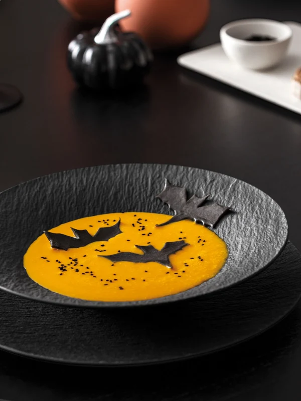 Un piatto nero con zuppa arancione e guarnizioni a forma di pipistrello su una tavola scura con un bicchiere da vino, posate nere e un piatto bianco con cibo sullo sfondo.