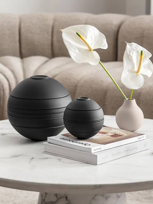 La Boule noire de Villeroy & Boch, un vase beige avec des fleurs blanches, un bougeoir et quelques livres dans une ambiance de salon beige.