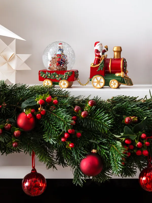 A festively decorated mantelpiece with the decorative train from the Christmas Toy collection by Villeroy & Boch. With red ornaments of a snow globe, star-shaped decorations and a green gift box decorated with a red bow and a candy cane.
