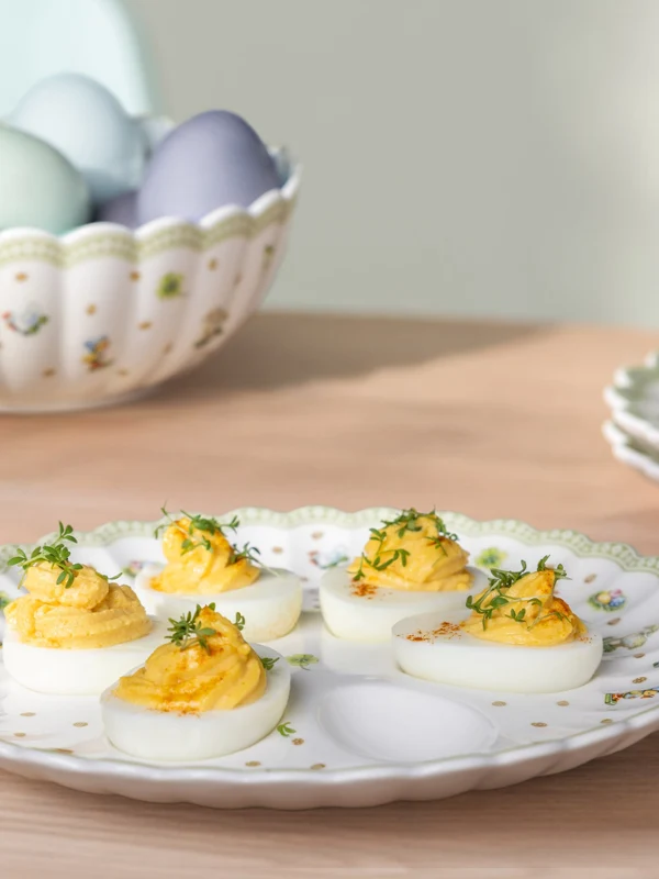 Ein Teller aus Easter Delight mit gefüllten Eiern, garniert mit Kräutern, steht auf einem Tisch neben einer Schale mit pastellfarbenen Eiern.