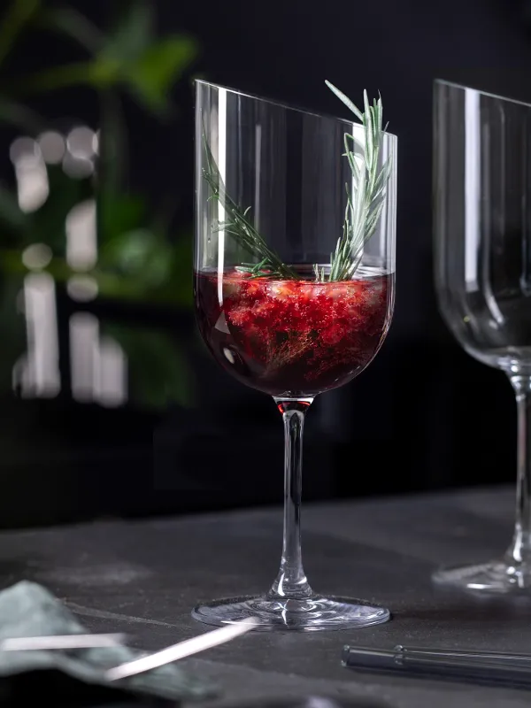 A Villeroy & Boch NewMoon glass filled with a red liquid decorated with fruit and a sprig of rosemary next to an empty glass on a dark table.