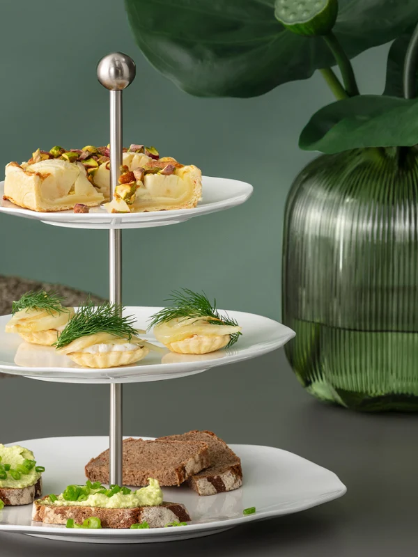 Three-tiered étagère from the Pura collection with various finger foods on a table, with green glass vases and plants in the background.