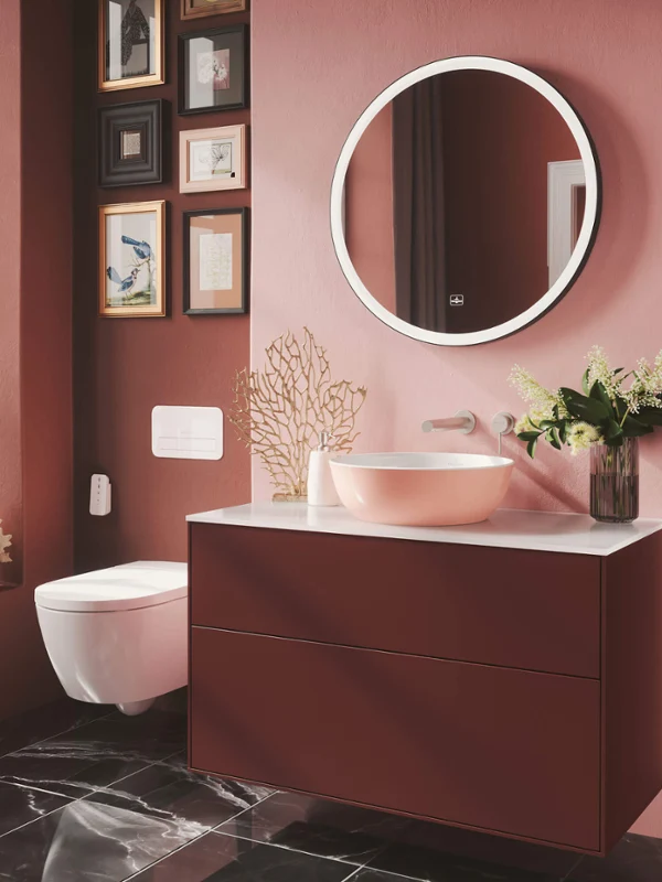 Różowa umywalka Artis na czerwonej szafce podumywalkowej Finion Nowoczesna łazienka z okrągłym lustrem, czerwoną szafką podumywalkową Finion i różową umywalką stojącą na blacie Artis, z ciemnoróżową ścianą i zdjęciami w ramkach