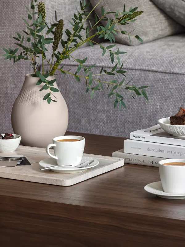 Un salon douillet avec une table basse en bois, sur laquelle se trouvent deux tasses à café Manufacture, un petit dessert, une pile de livres, une bougie et un vase Manufacture Collier avec des plantes vertes.