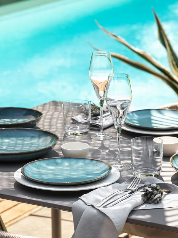 Das Tischset „Lave“ von Villeroy & Boch mit blauen Tellern, Gläsern und Besteck blickt auf einen türkisfarbenen Pool, während Pflanzenschatten Muster auf der Oberfläche erzeugen.