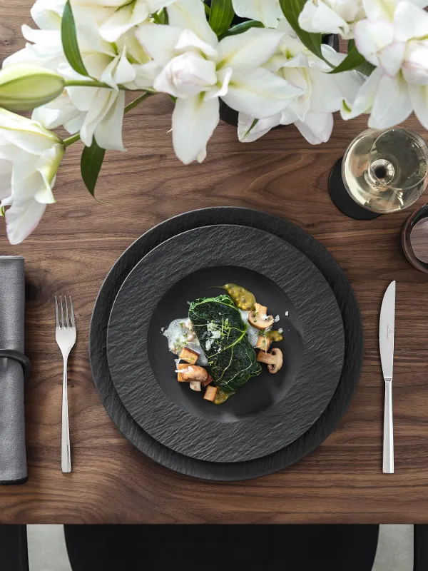 Mise en place élégante avec des assiettes Manufacture noires, des couverts, des figues, un plat de légumes et de crevettes, des serviettes grises, des fleurs blanches, un verre à vin et du pain coupé.