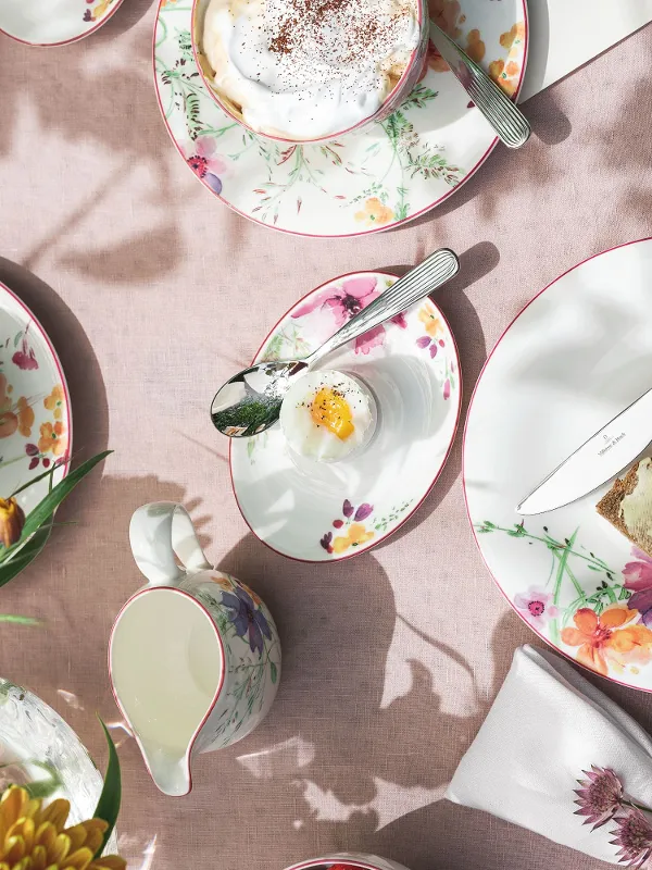 A Mariefleur place setting by Villeroy & Boch with toast and jam, an egg in a cup, and cappuccinos. A milk jug and flowers adorn the pink tablecloth.