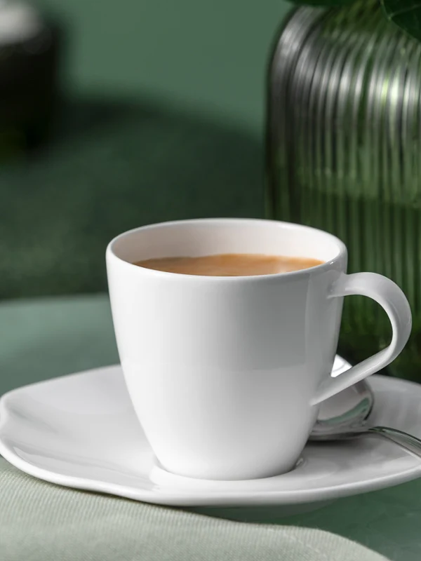 A white Pura coffee cup with saucer and spoon stands next to a green glass vase. A white Pura coffee cup with saucer and spoon stands on a green surface next to a green glass vase.