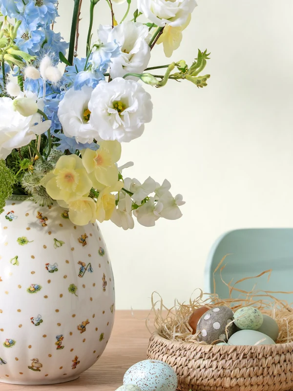 A Easter Delight vase with white and blue flowers sits beside a small nest filled with pastel eggs on a wooden table.