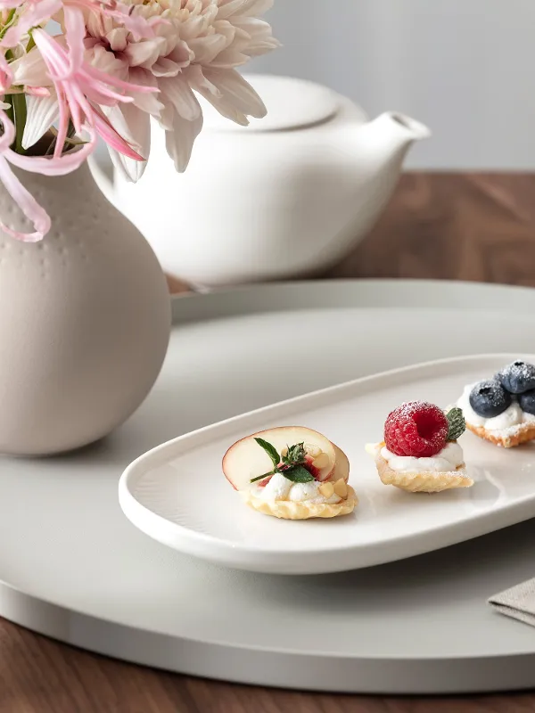 Un piatto bianco Afina con tre piccoli pasticcini e frutta si trova su un vassoio rotondo accanto a un vaso di fiori, una teiera e un brocchetto di latte su un tavolo di legno.
