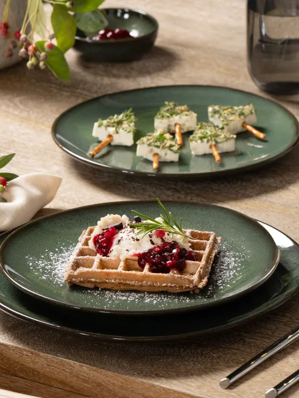 Une assiette verte Lave avec une gaufre à la crème et aux baies, une tasse de café Lave et des brochettes d'apéritifs sur une table en bois décorée pour les fêtes.