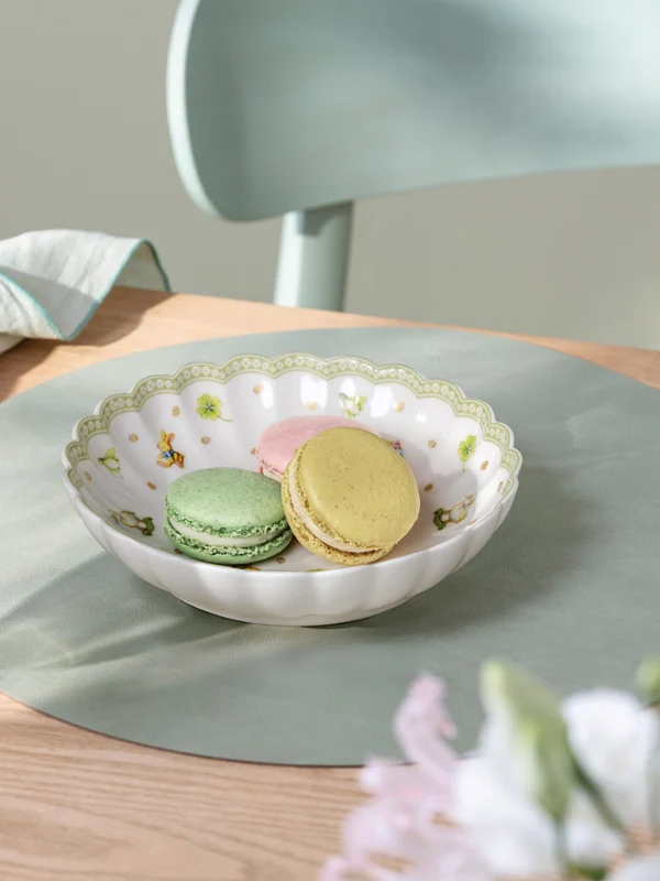 A Easter Delight bowl sits on a light green Easter Delight placemat A Easter Delight bowl of pastel macarons sits on a light green Easter Delight placemat beside a glass and a folded napkin on a wooden table.