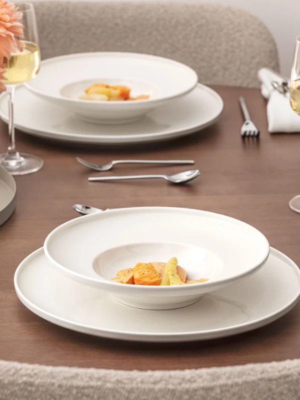 A neatly laid dining table from the Afina collection with two plates of gourmet food, wine glasses, water glasses, silver cutlery, cloth napkins and a vase of peach blossoms on a wooden surface.