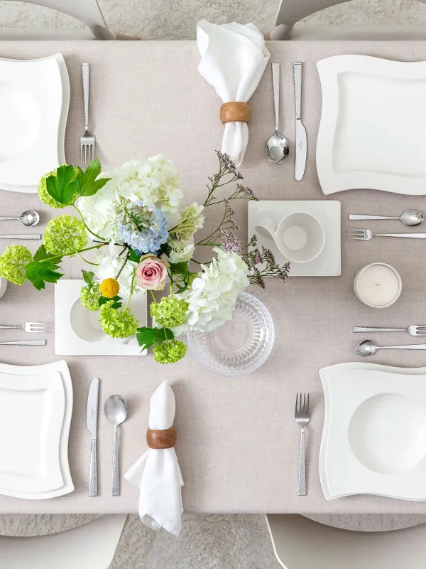 Apparecchiatura elegante con piatti quadrati bianchi, anelli per tovaglioli, posate disposte ordinatamente, centrotavola floreale con fiori bianchi e verdi e una brocca di vetro con fette di limone.