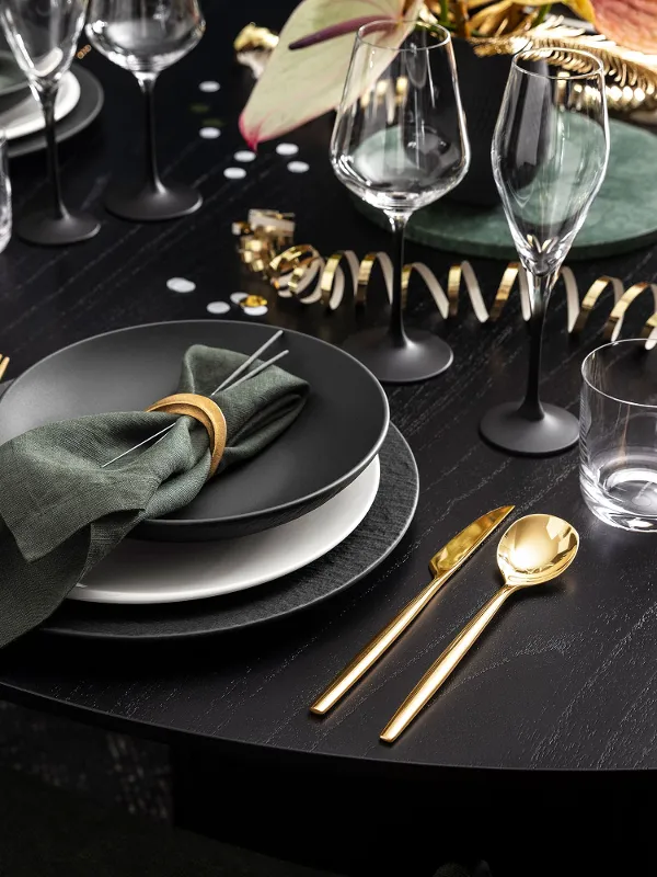 Une élégante décoration de table pour le Nouvel An avec des assiettes Manufacture noires et des couverts de table dorés sur une table ronde sombre, décorée d’une composition florale et de confettis festifs.