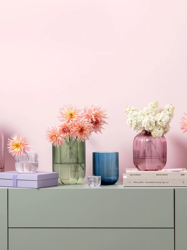 Fleur glass vases in various pastel colors and sizes Various glass vases in pastel colors from the Fleur collection, decorated with flowers, are arranged on a light surface with books and small decorative items in front of a pink wall.