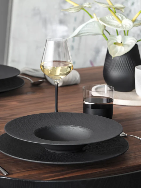 Ein modernes Esszimmer-Set aus Holz mit Geschirr von Manufacture, Weingläsern, Besteck und einer Vase mit weißen Blumen.