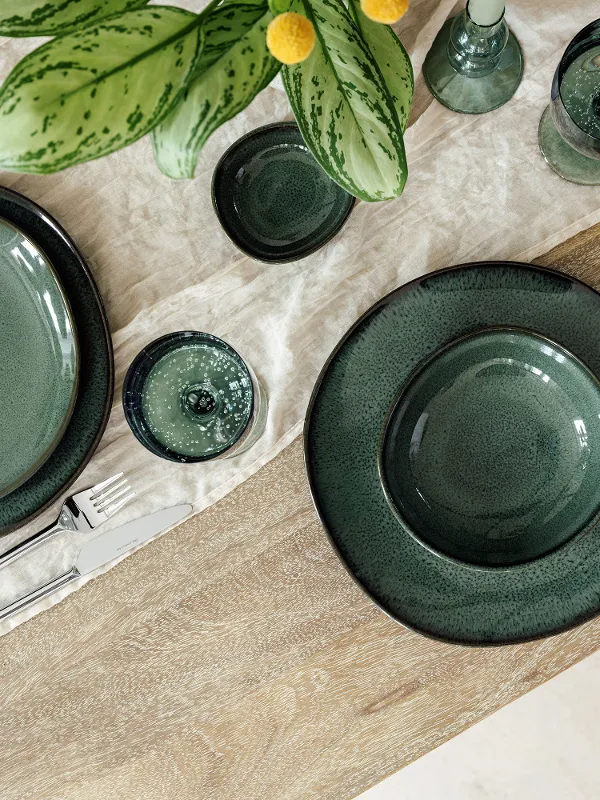Table en bois élégamment dressée avec de la vaisselle en céramique "Lave Vert", des couverts en argent "like" de like. by Villeroy & Boch et des verres Like assortis sur une serviette en tissu crème. Une plante verte avec des accents jaunes complète l'arrangement raffiné.