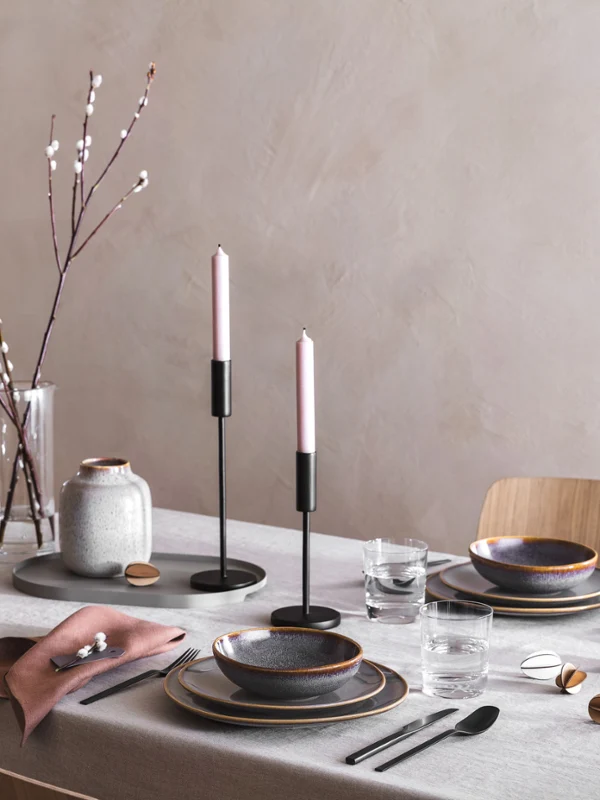 An elegant dining table with lit candles, Lave ceramic crockery, black Manufacture cutlery and minimalist Easter decor with willow catkins.