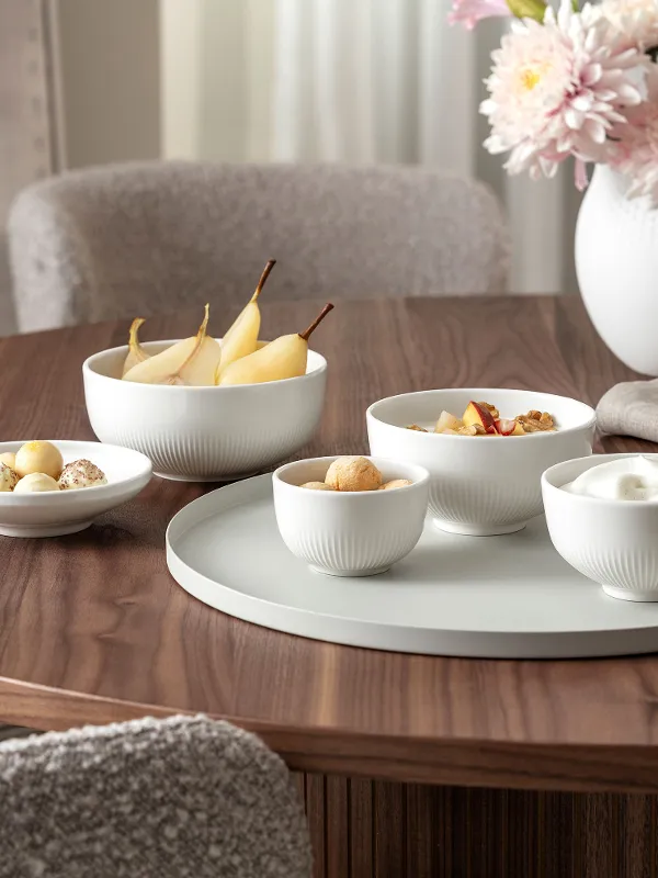 Une table ronde en bois avec des bols Afina remplis de snacks, de poires et un vase de fleurs soigneusement disposés dessus.