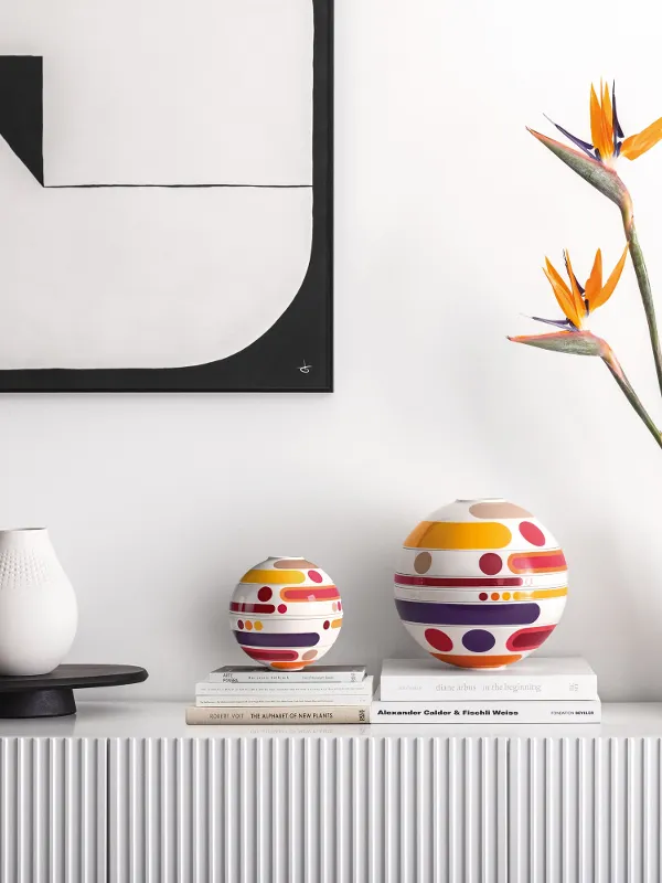 A minimalist sideboard showcases the modern La Boule Miami, vases, bird of paradise flowers, and stacked books under abstract wall art.