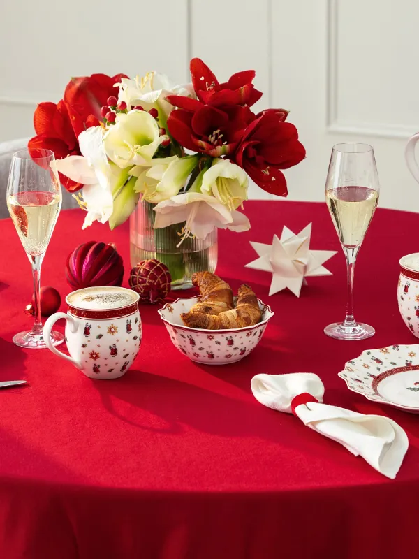 Tavolo rotondo con una tovaglia rossa apparecchiata per l'ora del tè, con porcellana festiva, pasticcini, fiori e decorazioni natalizie.