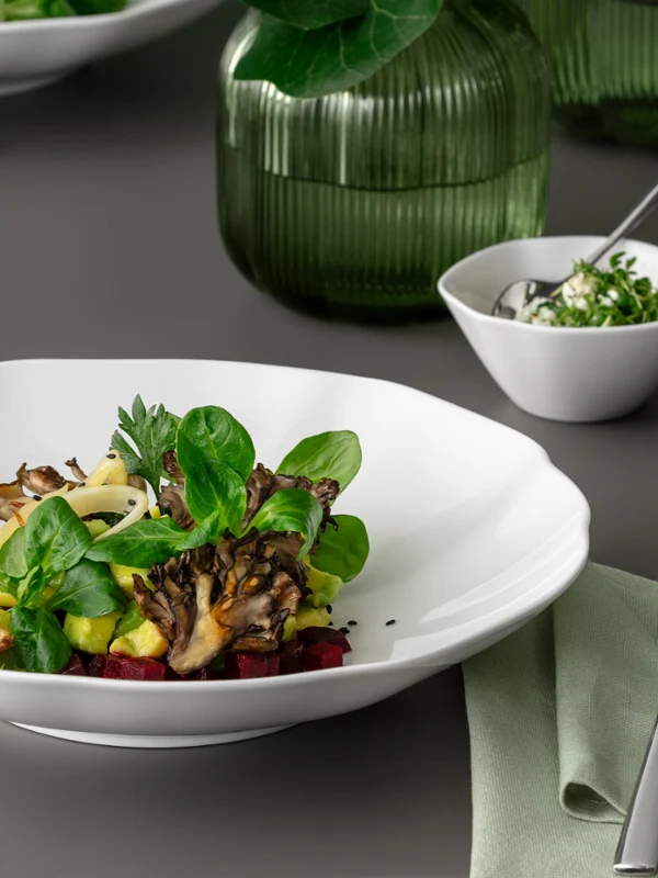 A white Pura bowl with a salad of greens and vegetables on a grey table, next to it cutlery and a small bowl, also from the Pura collection, with herbs.