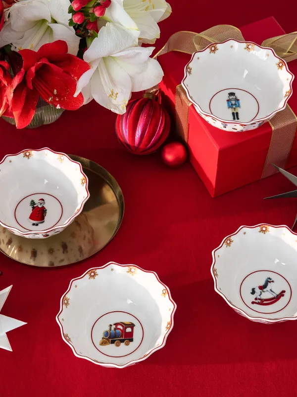 Quattro ciotole decorative di Christmas Toys Memory con motivi natalizi su una tovaglia rossa, circondate da ornamenti, fiori e una confezione regalo.