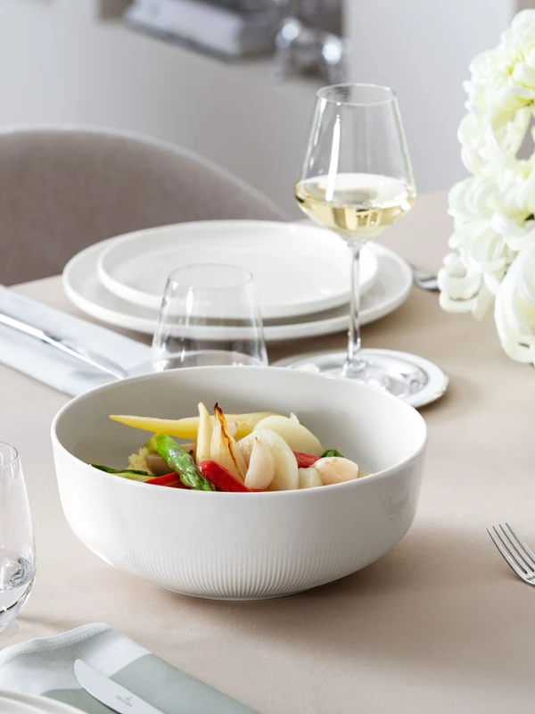 Un grand plat creux de service Afina de Villeroy & Boch, rempli de légumes colorés, sur une table dressée avec des fleurs en arrière-plan.