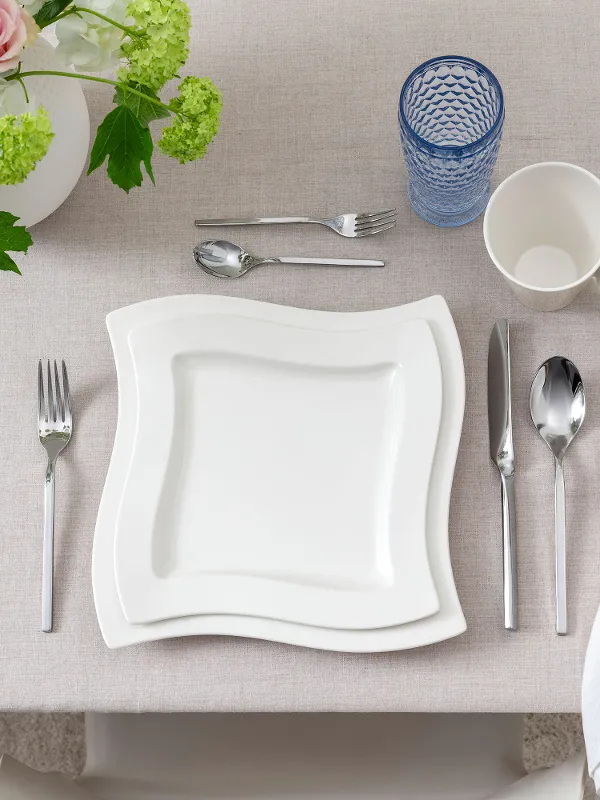 Clean, modern top view of a single table setting featuring NewWave porcelain plates and cup, Boston Blue long drink glass, and matching NewWave cutlery. Neutral tones and minimalist aesthetic create a contemporary dining impression.