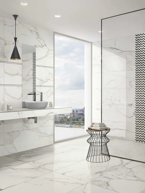 Nowoczesna łazienka z umywalką podwójną Artis firmy Villeroy & Boch Nowoczesna łazienka z białymi marmurowymi powierzchniami, umywalką podwójną Artis firmy Villeroy & Boch, czarnymi lampami wiszącymi, brodzikiem bezprogowym z kafelkami w geometryczne wzory, stolikiem z drucianym koszem i oknem z widokiem na miasto.