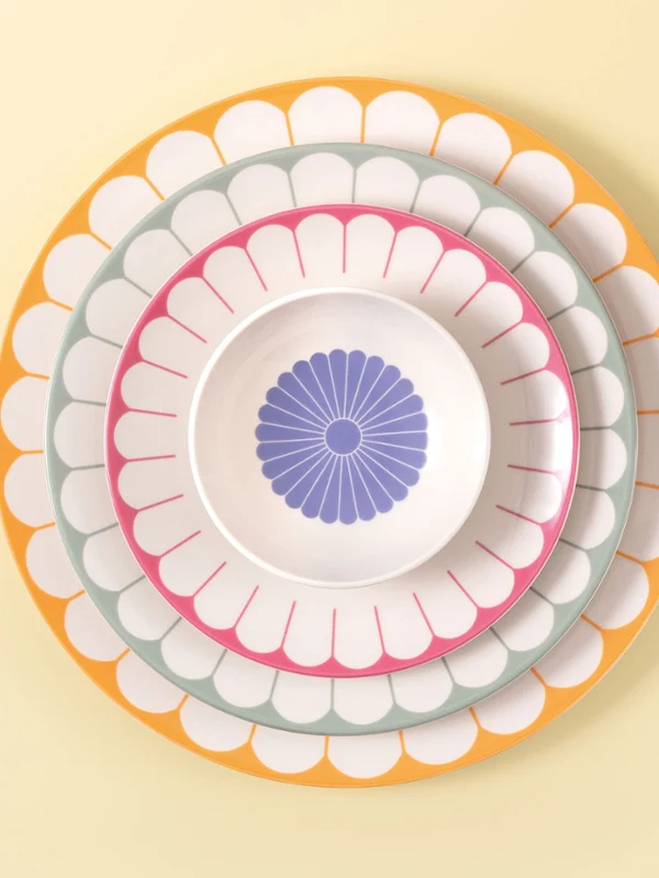 Four Villeroy & Boch Fleur plates in different sizes and colours placed on a light yellow background next to pink flowers.