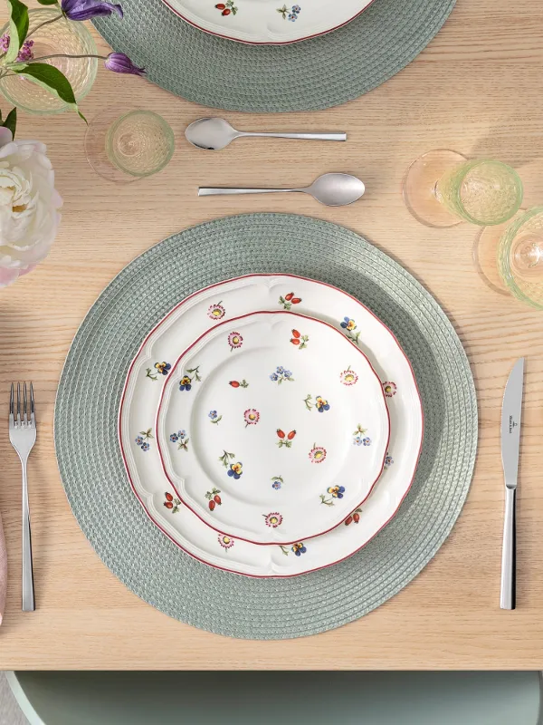 A set table with Petite Fleur flower plates, cutlery, a pink napkin, a green placemat, and a bouquet of flowers on a wooden table. 