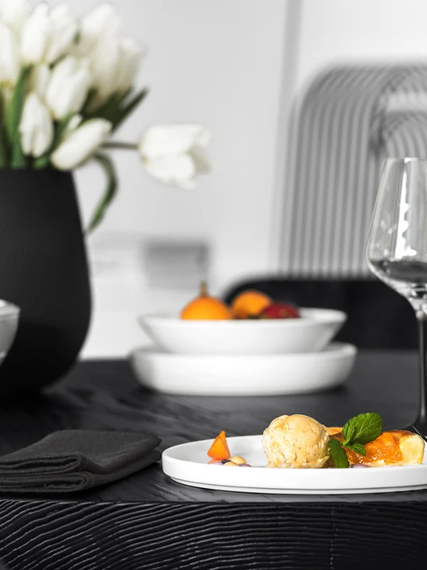 Dessert on white La Boule A dark set table with a dessert, two wine glasses, a white La Boule bowl with fruit, and a black Collier vase with white tulips.