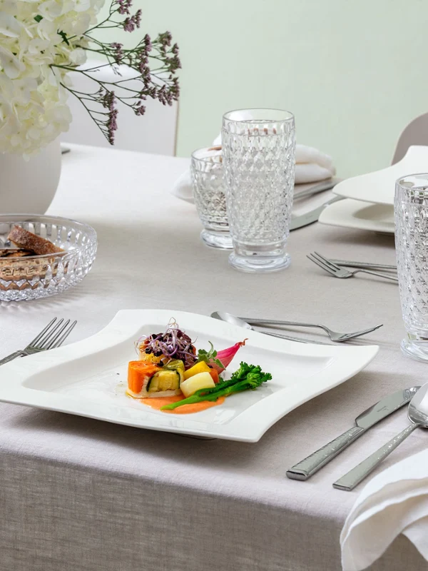 Side view of a beautifully set dining table featuring the NewWave dinner plate with a refined vegetable-based dish. Accompanied by Boston and Boston Green tumblers and Blacksmith cutlery. A stylish and elegant setup for a gourmet meal.